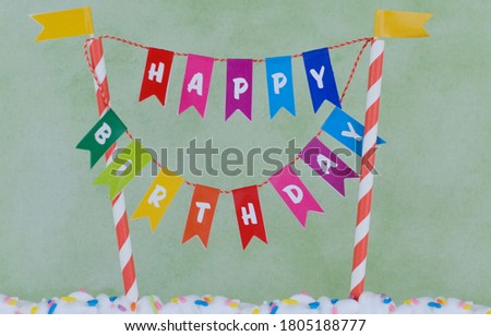Cake topper bunting on striped posts spelling Happy Birthday. Rainbow sprinkles decorate the white icing. Background is sponged green.