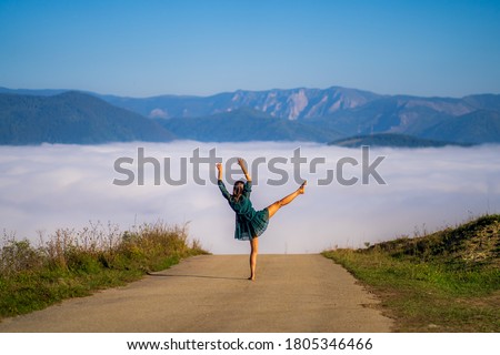 Beautiful Caucasian woman dressed in an elegant green dress dancing barefoot on a high road leading to the distant mountains covered with fog during a sunny day in Romania. Royalty-Free Stock Photo #1805346466