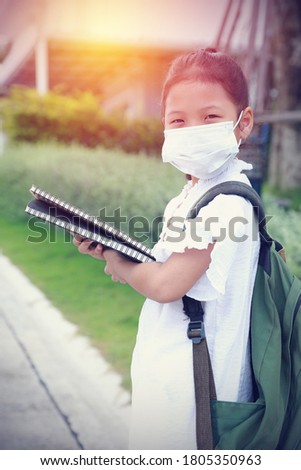 Asian little kid girl wearing the mask medical holding books and carry the green backpack looking at the camera. Back to school because there is a big outbreak of the coronavirus.