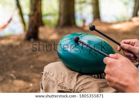 Close-up with the hands of a Caucasian musician holding drum sticks and playing a modern hand pan steel tongue drum percussion instrument in the middle of nature. Royalty-Free Stock Photo #1805355871