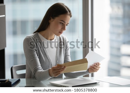 Serious confident businesswoman reading letter, holding open envelope, sitting at desk in modern office, received news or important information, young woman working with correspondence Royalty-Free Stock Photo #1805378800