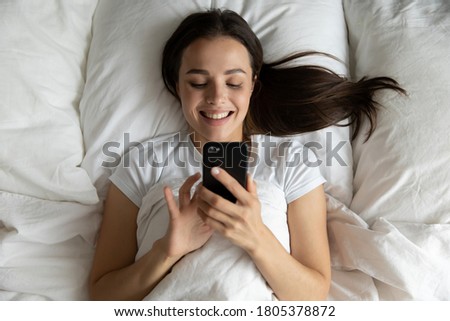 Top view smiling young woman using phone, relaxing in bed, lying on soft pillow under warm duvet, checking social networks after awakening, happy beautiful girl looking at smartphone screen