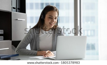 Smiling young woman employee wearing headset writing notes, studying online, sitting at desk in modern office, watching webinar or training, listening to lecture, looking at laptop screen Royalty-Free Stock Photo #1805378983