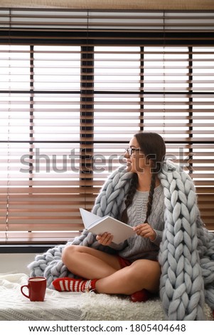 Happy young woman wrapped in knitted blanket with book near window at home. Lazy morning