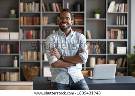 Portrait smiling African American businessman standing near work desk in modern office room, successful confident young man wearing glasses posing for photo with arms crossed, looking at camera Royalty-Free Stock Photo #1805431948
