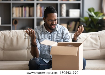 Angry irritated African American man in glasses dissatisfied by received parcel, unhappy customer unpacking cardboard box, wrong or broken fragile online store order, bad delivery service concept Royalty-Free Stock Photo #1805431990