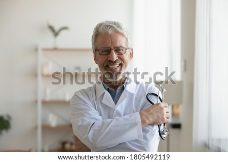Head shot portrait smiling mature doctor holding stethoscope looking at camera, happy confident senior therapist gp wearing white uniform standing in modern office, healthcare and insurance concept