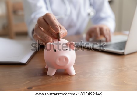 Close up young woman doctor putting coin into pink piggy bank, sitting at work desk, using laptop, hospital budget and accounting, finances, medical insurance, healthcare money savings concept