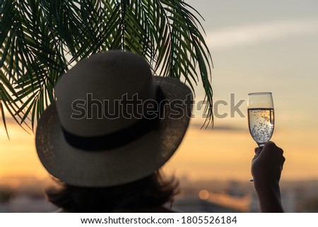 Back view of lonely beautiful woman with glass of champagn  in a straw hat against the background of the sea in branches of palm trees. Sunset beach. Summer and freedom concept.