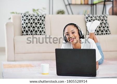 Portrait of positive young Black woman wearing headphones when lying on the floor and working on laptop at home