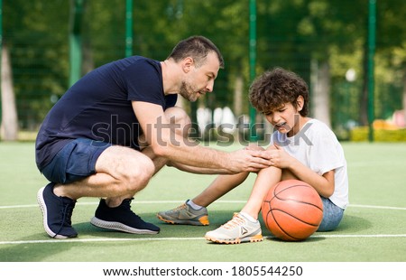 Sports Injury. Handsome PE teacher helping boy with knee trauma after playing basketball Royalty-Free Stock Photo #1805544250