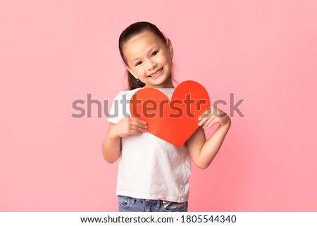 Kindness Concept. Portrait of adorable asian pupil holding big red paper heart over pink studio wall, copy space