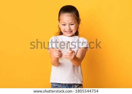 Cute asian child using smart phone and smiling, standing over pastel yellow wall, copyspace