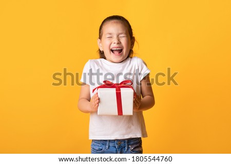 Wow. Excited little kid holding gift box and screaming with joy, closed eyes and open mouth, yellow wall, copyspace