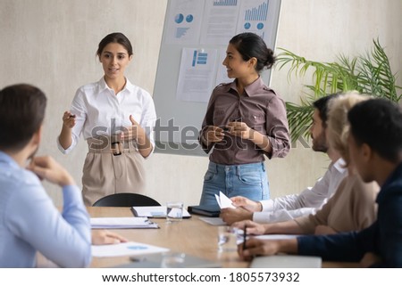 Skilled young arab female presenter standing near flipchart with happy indian colleague, presenting new marketing strategy to focused mixed race business people investors at briefing meeting in office Royalty-Free Stock Photo #1805573932