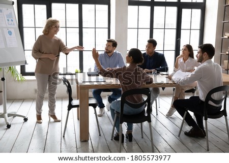 Interested young indian woman raising hand, asking questions to older pleasant female speaker leader coach trainer on educational lecture seminar workshop with mixed race colleagues in office. Royalty-Free Stock Photo #1805573977