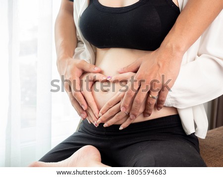 portrait of 4 months pregnant Asian woman in white bed room, Woman touching her abdomen belly on white background. male and female hands in heart shape.