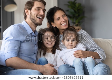 Smiling young married couple relaxing on sofa with adorable little children daughters, enjoying watching TV program, kids show or cartoons together at home, happy family hobby weekend pastime. Royalty-Free Stock Photo #1805623399