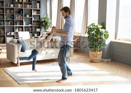 Full length playful young father holding hands of energetic small child daughter, swaying baby girl in air, entertaining together in modern living room, happy family weekend holiday pastime concept. Royalty-Free Stock Photo #1805623417