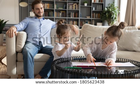 Interested engaged adorable preschool children sisters drawing pictures in paper album on coffee table while handsome young father resting on comfortable couch, weekend daycare family pastime.