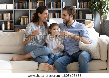 Happy young couple parents teaching little preschool daughter drinking clear water every day. Smiling healthy family holding glasses with pure aqua, enjoying morning daily healthcare habit at home. Royalty-Free Stock Photo #1805623636