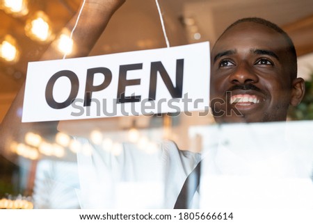 Happy young African waiter with toothy smile hanging paper with notice open on transparent door of cafe or restaurant in the morning