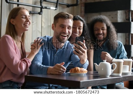 Overjoyed diverse young people posing for photo in cafe together, smiling excited man holding smartphone, taking selfie or recording video for social network, having fun with friends
