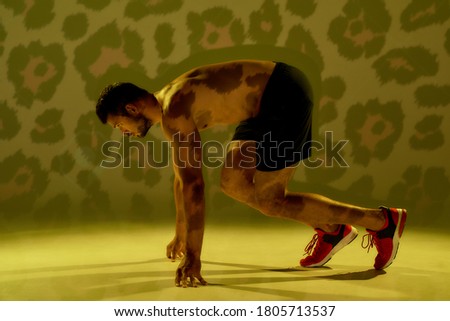 Run like a devil. Side view of a young muscular caucasian man standing in start position, ready for run isolated over yellow cheetah background. Running, jogging, workout. Healthy lifestyle and sport