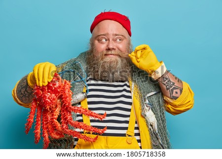 Cheerful seaman curls mustache, holds big sea creature, dressed in striped jumper and yellow overalls, travels by sea, enjoys summer travel, poses indoor, blue background. Yachting, adventure concept