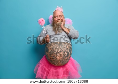 Dreamy man with thick beard, big belly, takes part in cartoon performance, has happy upbringing memories, wears fairy outfit, holds magic wand, plays with daughter, partying together, poses indoor