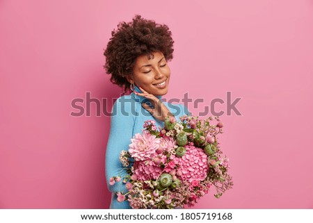 Charming young woman with gentle smile, touches face, stands indoor, keeps eyes closed, receives bouquet of flowers from colleagues in honor of her promotion, models against pink background.