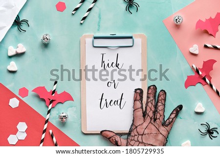 Trick or treat text on clipboard pad on layered paper background in mint green and red. Flat lay, Halloween party, hand in glove, confetti, drink straws, bat silhouettes, sugar hearts and spiders.