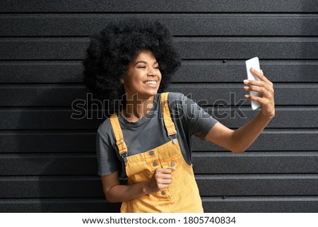 Happy African American hipster woman blogger with Afro hair laughing, holding phone, recording vlog, shooting social media stories or video calling in mobile app standing on black background. Royalty-Free Stock Photo #1805740834