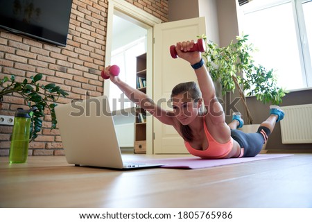 Active fit sporty young woman holding dumbbells doing fitness exercise training abs and back at home on mat watching online video training tv tutorial sport live stream workout class on laptop. Royalty-Free Stock Photo #1805765986