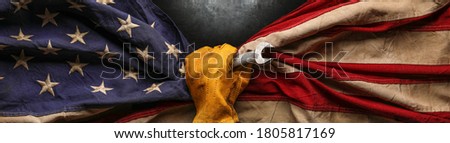 Worn work glove holding wrench tool and gripping old worn US American flag. Made in USA, American workforce, blue collar worker, or Labor Day concept. Royalty-Free Stock Photo #1805817169