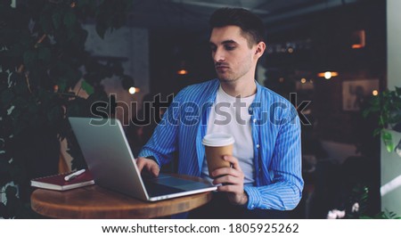 Caucasian male IT professional checking program text on modern laptop computer doing distance job in coffee shop, handsome man with takeaway cup watching online web webinar via application platform