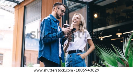 Cheerful caucasian female laughing at funny media content of boyfriend's mobile phone spending free time together, male holding mobile phone showing video from social network to woman collegue