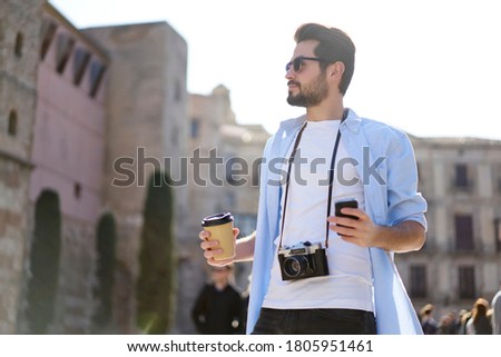 Low angle of serious bearded male tourist in casual wear and sunglasses with camera looking away while drinking coffee and surfing smartphone