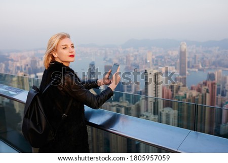 Side view of content female tourist in casual clothes with backpack standing on rooftop of contemporary building in downtown of Hong Kong and looking away while taking photo on mobile phone
