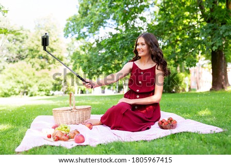 leisure and people concept - happy smiling woman with smartphone on selfie stick and picnic basket sitting on blanket and taking picture at summer park
