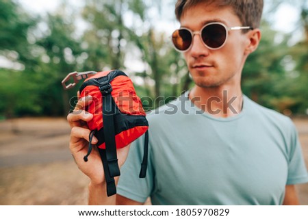 Close photo of a young man in sunglasses holding in his hand a little red bag of camping equipment on a background of forest. Active rest in the mountains. Bag hammock for tourism.