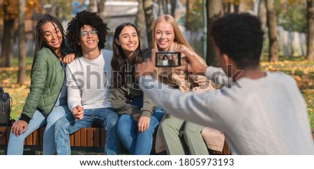 Black guy taking photo of his international friends in park, using smartphone, panorama