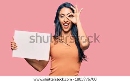 Beautiful hispanic woman holding blank empty banner smiling happy doing ok sign with hand on eye looking through fingers 