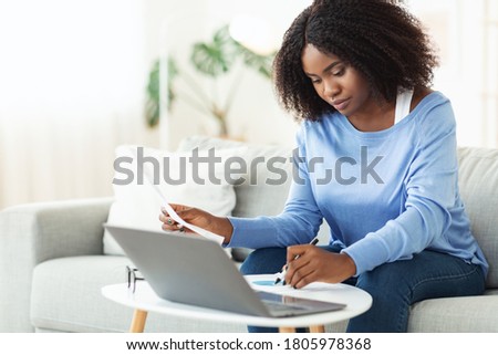 Portrait of professional black woman at work, reading and writing some papers sitting on couch at table with pc Royalty-Free Stock Photo #1805978368