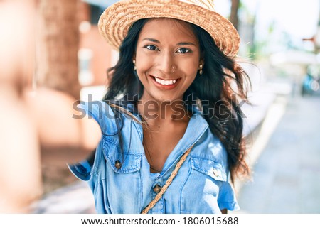 Young beautiful indian woman wearing summer hat smiling happy making selfie by the camera at the city.