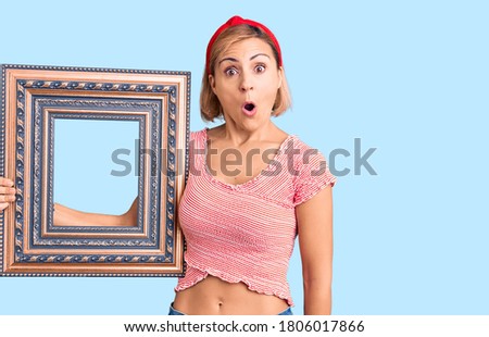Young blonde woman holding empty frame scared and amazed with open mouth for surprise, disbelief face 