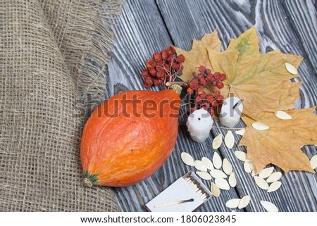 Orange pumpkin, seeds and candle stubs. Nearby are a box of matches, dried maple leaves and rowan. On brushed pine boards painted black and white.