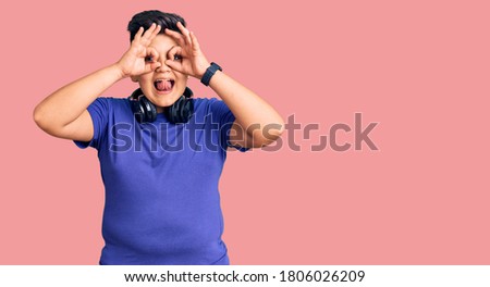 Little boy kid listening to music wearing headphones doing ok gesture like binoculars sticking tongue out, eyes looking through fingers. crazy expression. 