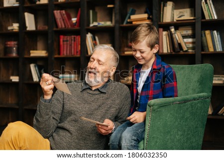 Senior bearded gray-haired grandfather with his grandson enjoy memories watching family photo album sitting on armchair and floor at home in living room with an authentic aristocratic interior.