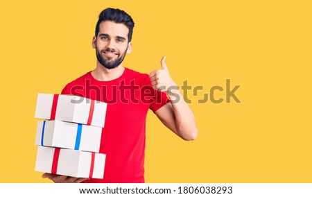 Young handsome man with beard holding gift smiling happy and positive, thumb up doing excellent and approval sign 